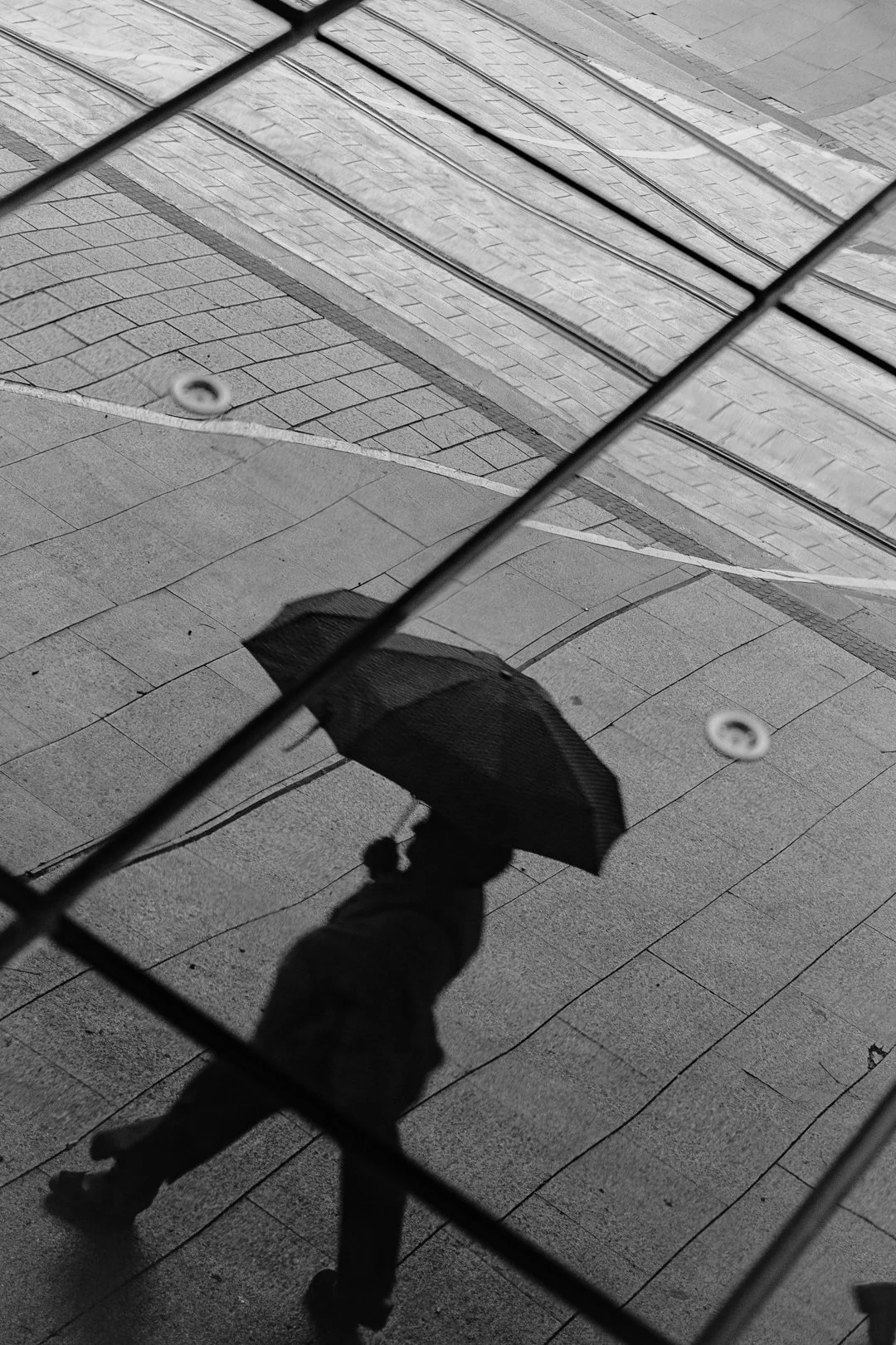 Black-and-white shadow of a person holding an umbrella on a tiled sidewalk, viewed through a chain-link fence
