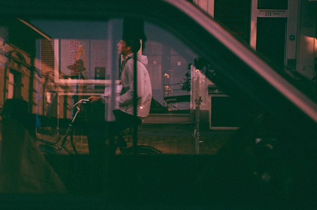 Man riding a bicycle past a storefront at night, viewed through a car window