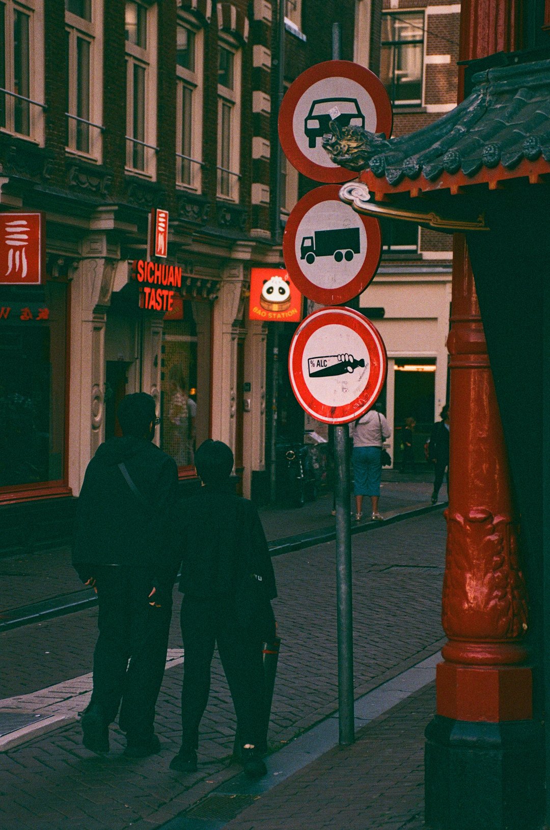 Street scene with stacked traffic signs, a Chinese restaurant, ornate architecture, and pedestrians on a cobblestone walkway