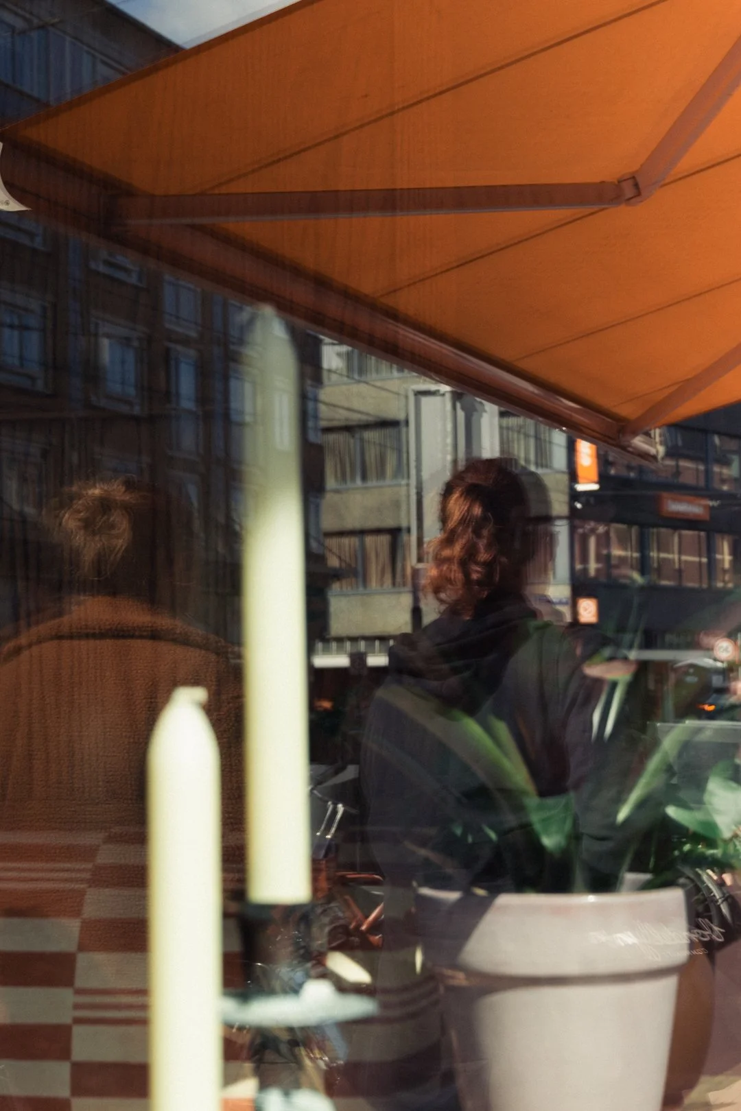 People reflected in a cafe window beneath an orange awning, with a potted plant and city street outside.