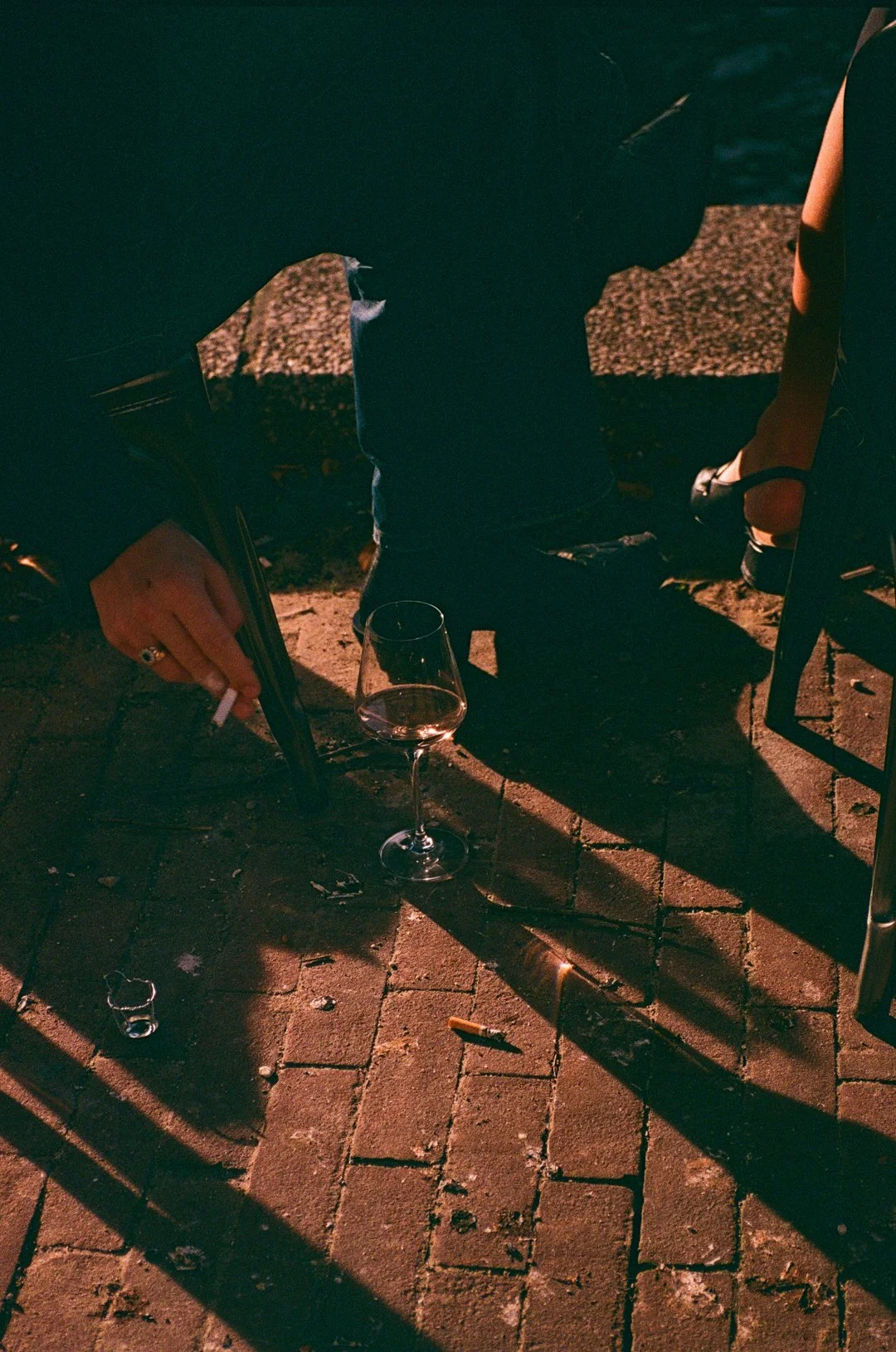 Low-lit outdoor scene with a person holding a cigarette beside a half-filled wine glass on a brick patio.