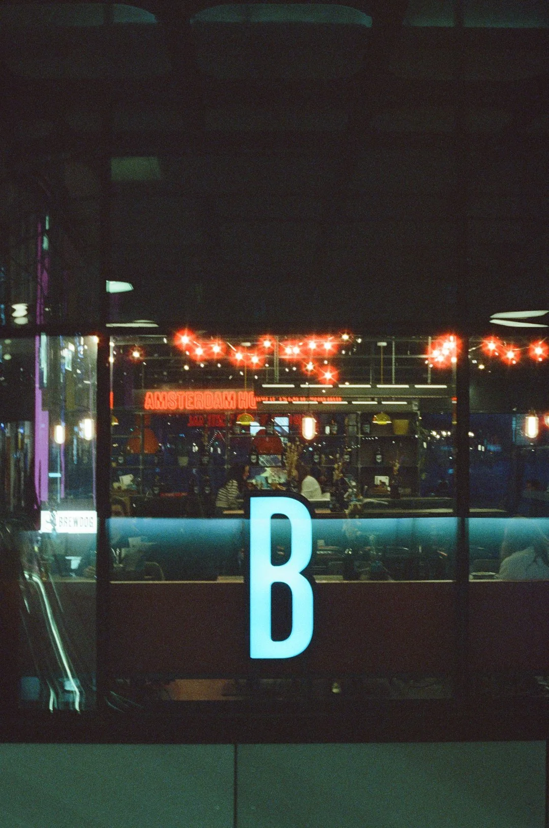 Nighttime view of a brightly lit Amsterdam-themed bar or restaurant, with a large illuminated letter B in the foreground.