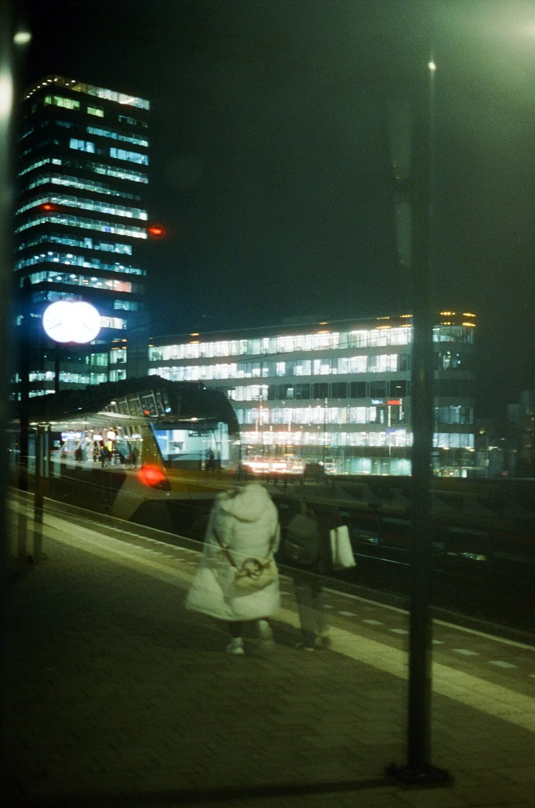 Nighttime city scene with illuminated office buildings, a large clock, and pedestrians on a station platform.