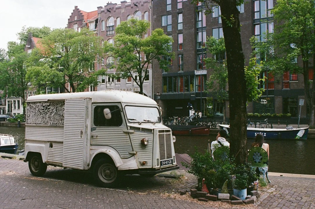Vintage white delivery truck parked beside a tree-lined canal in Amsterdam, with boats and historic buildings in the background