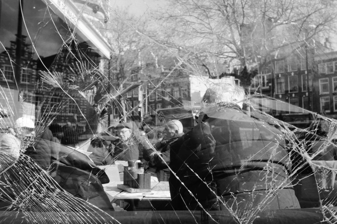 Black-and-white photo of people dining inside a restaurant, viewed through a heavily cracked window with a city street reflected in the glass.