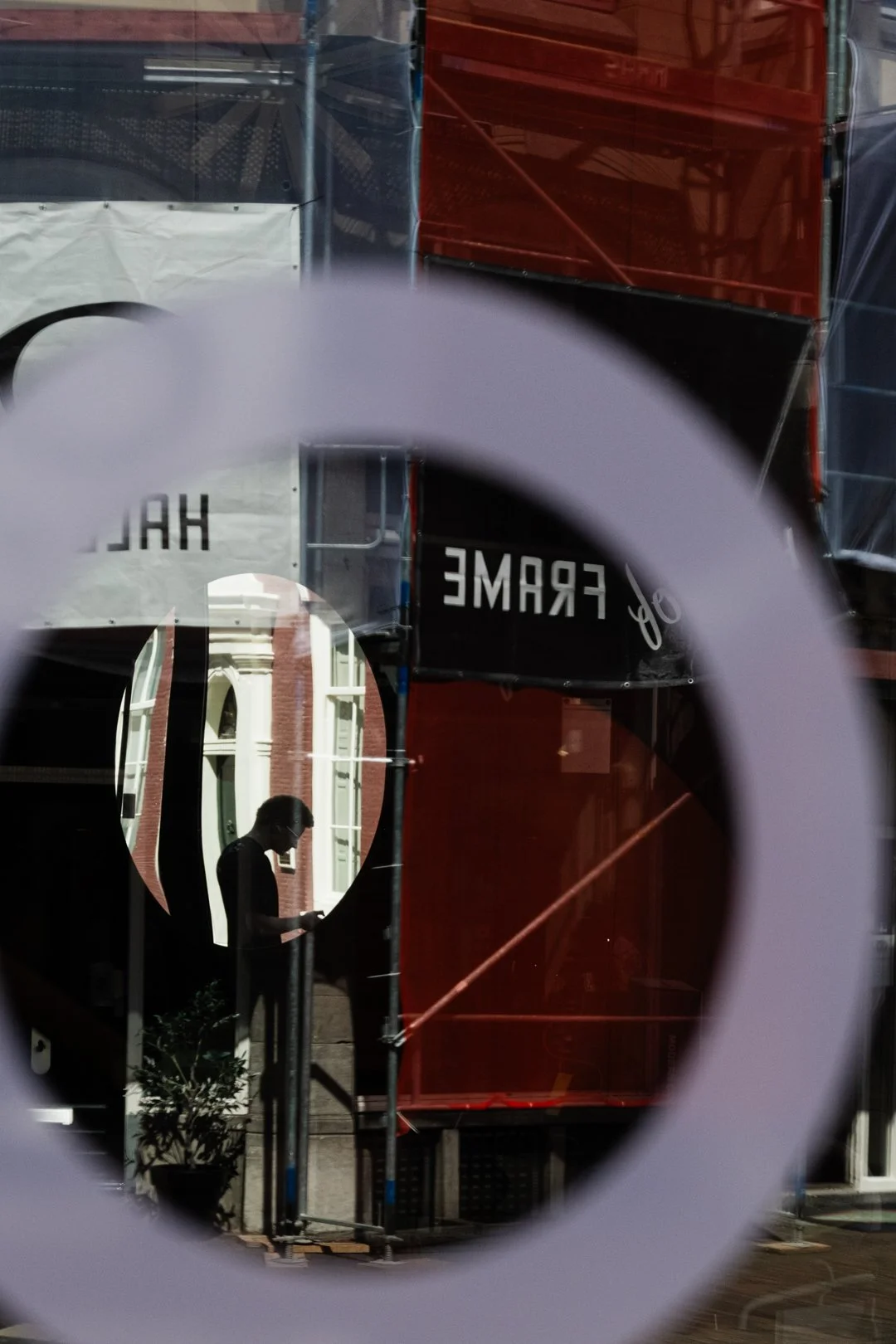 City street reflected in a shop window, with red construction scaffolding, reversed “FRAME” lettering, and a silhouetted person.