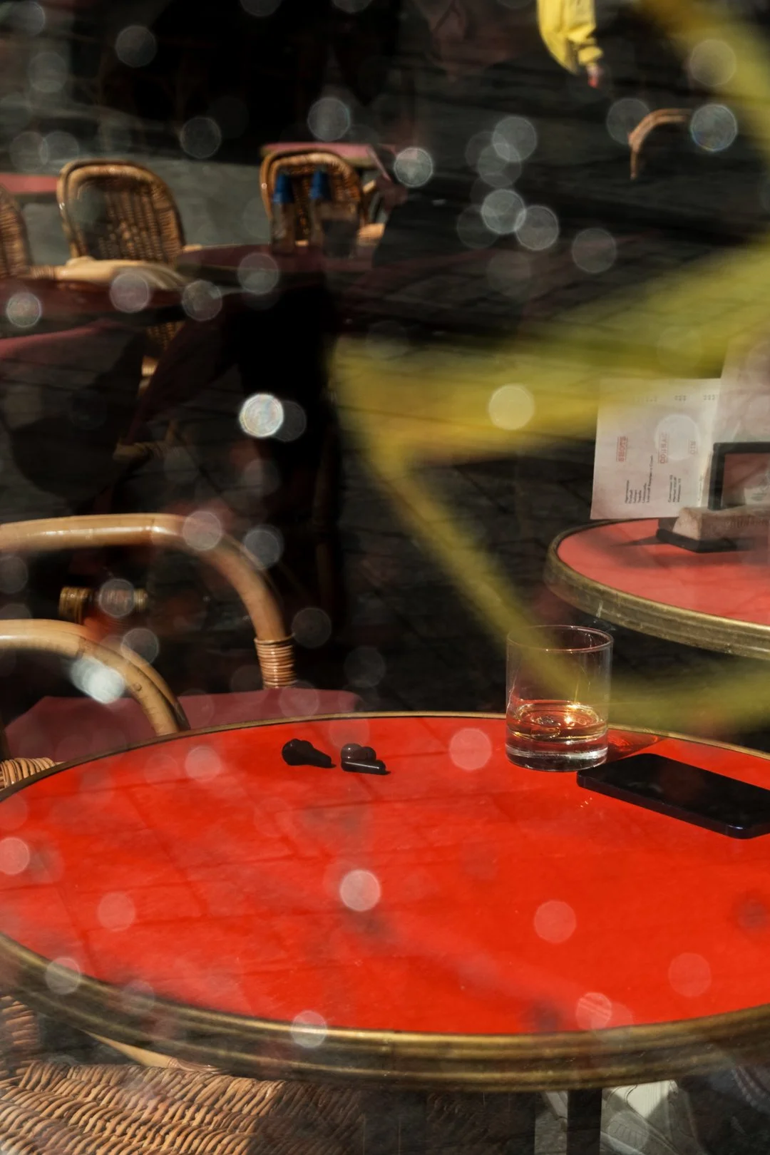 Red café table with a whiskey glass and smartphone, surrounded by wicker chairs and blurred reflections.