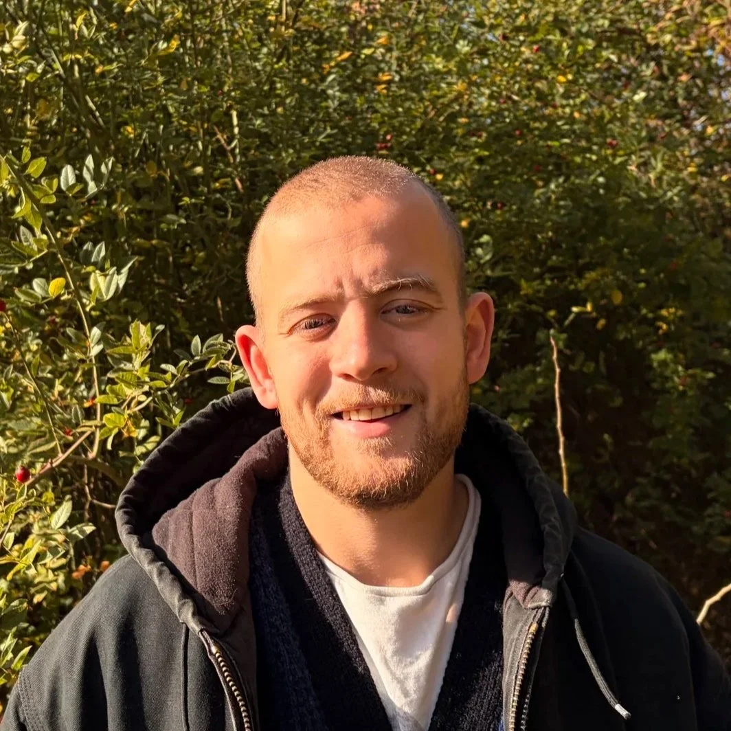 Smiling bald man in a black hooded jacket posing outdoors in front of leafy bushes