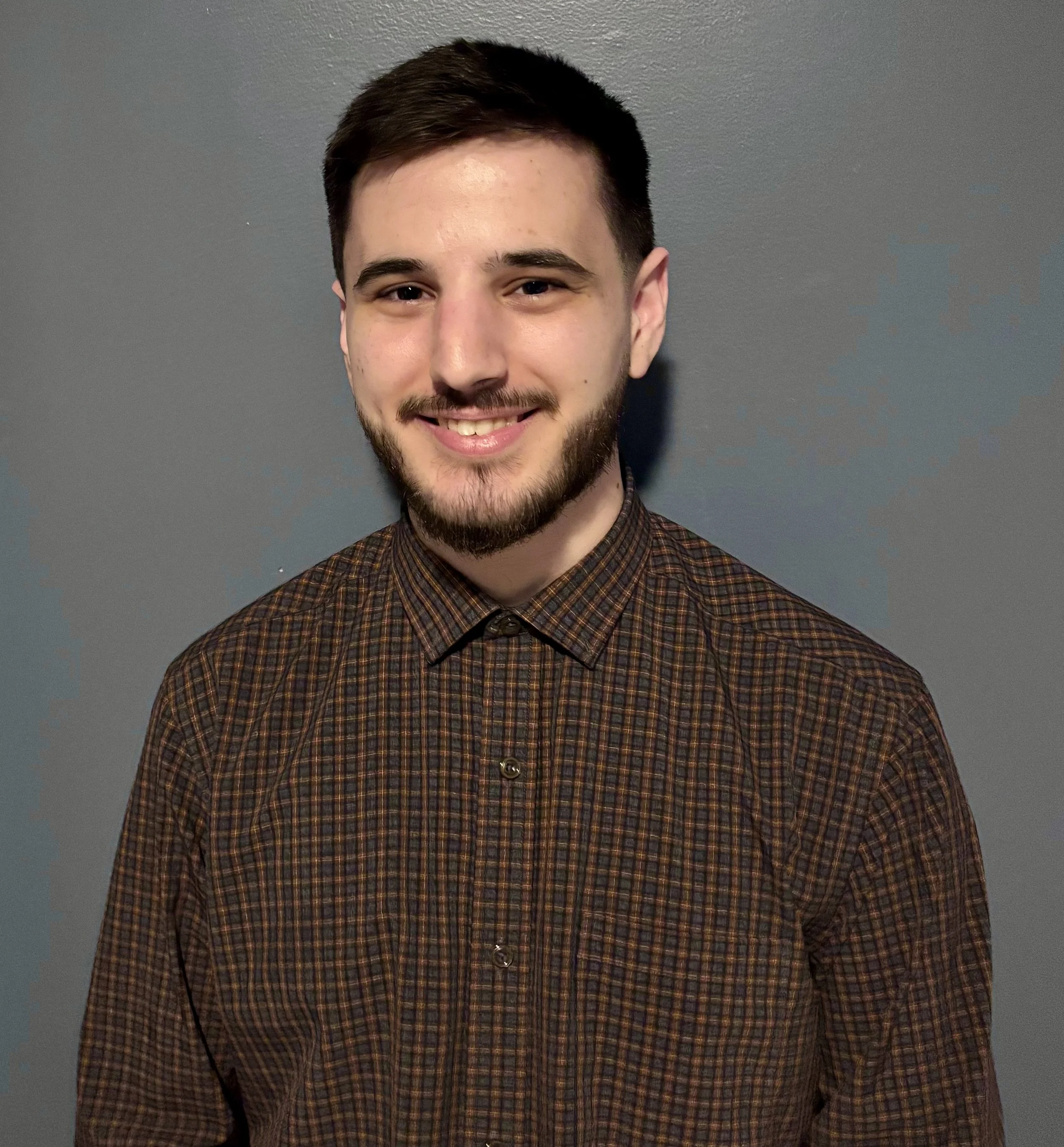 Smiling bearded man wearing a brown plaid button-down shirt against a gray wall