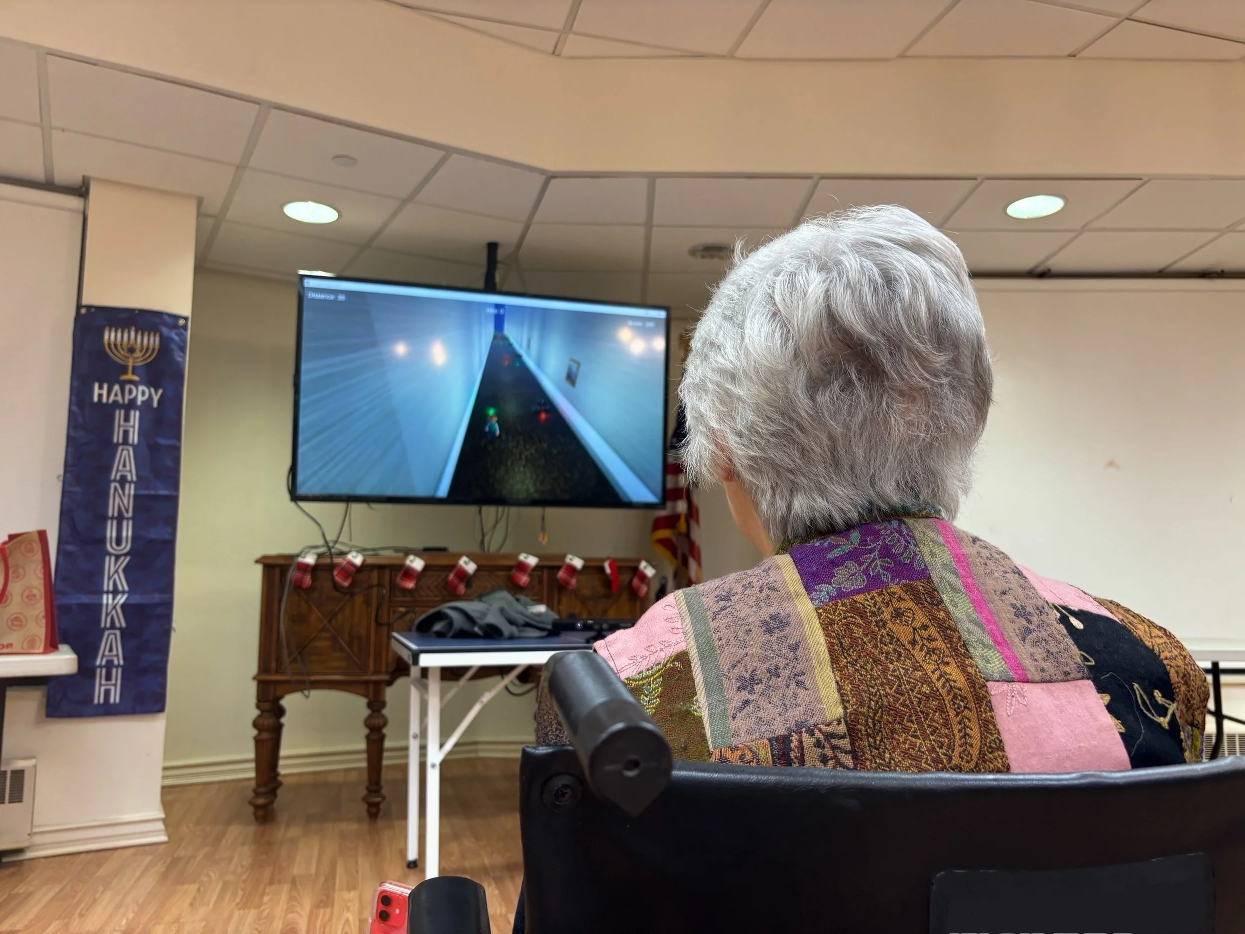 Older woman in a wheelchair watching a video game on a wall-mounted TV in a decorated community room with Hanukkah and Christmas decorations.