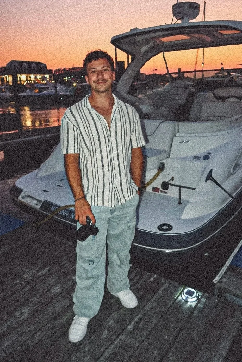 Man holding a camera stands on a dock beside a motorboat at sunset in a marina.