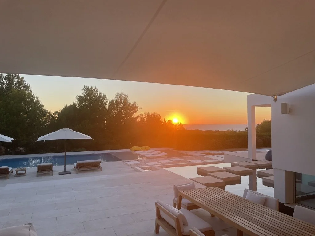 Sunset over the ocean viewed from a modern poolside patio with lounge chairs, umbrellas, and outdoor dining furniture