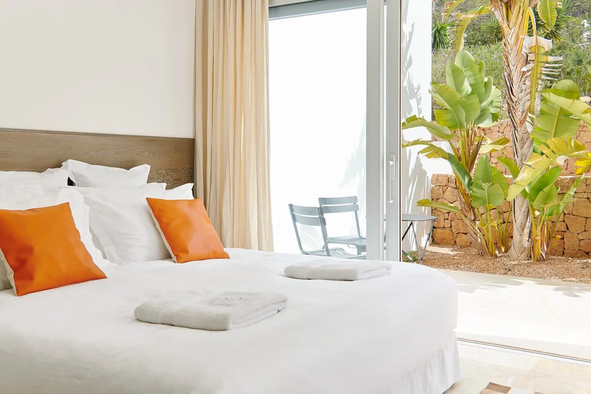 Bright modern bedroom with white bed, orange pillows, folded towels, and a glass door overlooking tropical plants and a stone wall