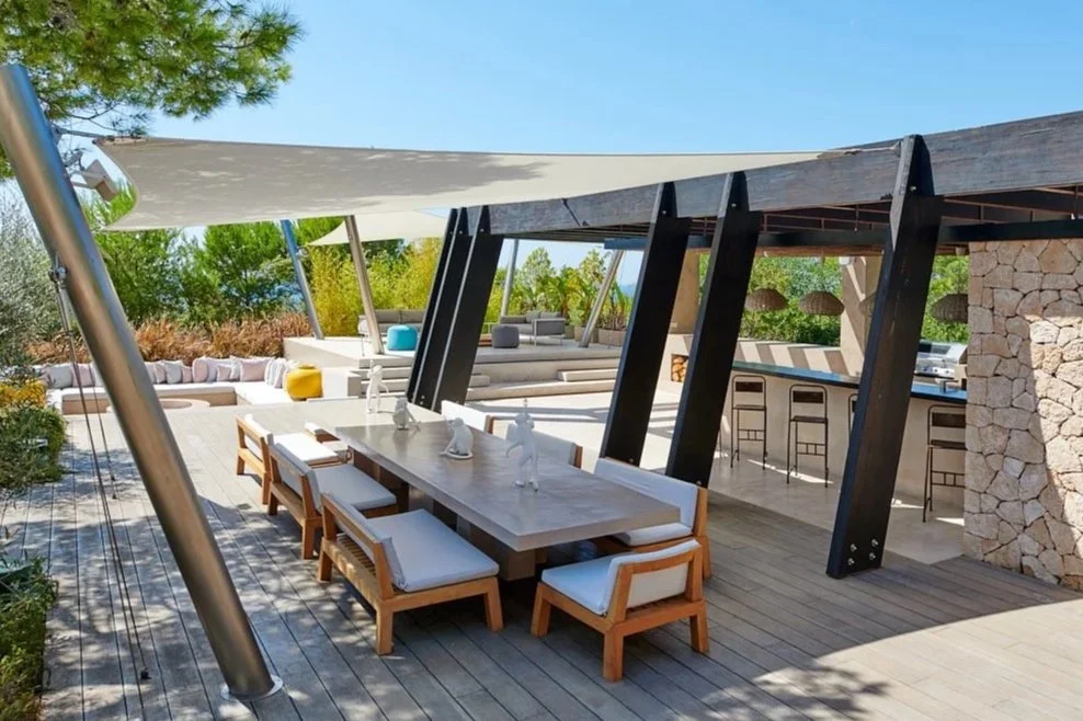 Modern outdoor terrace with dining table, lounge seating, bar, stone wall, and black structural beams surrounded by trees