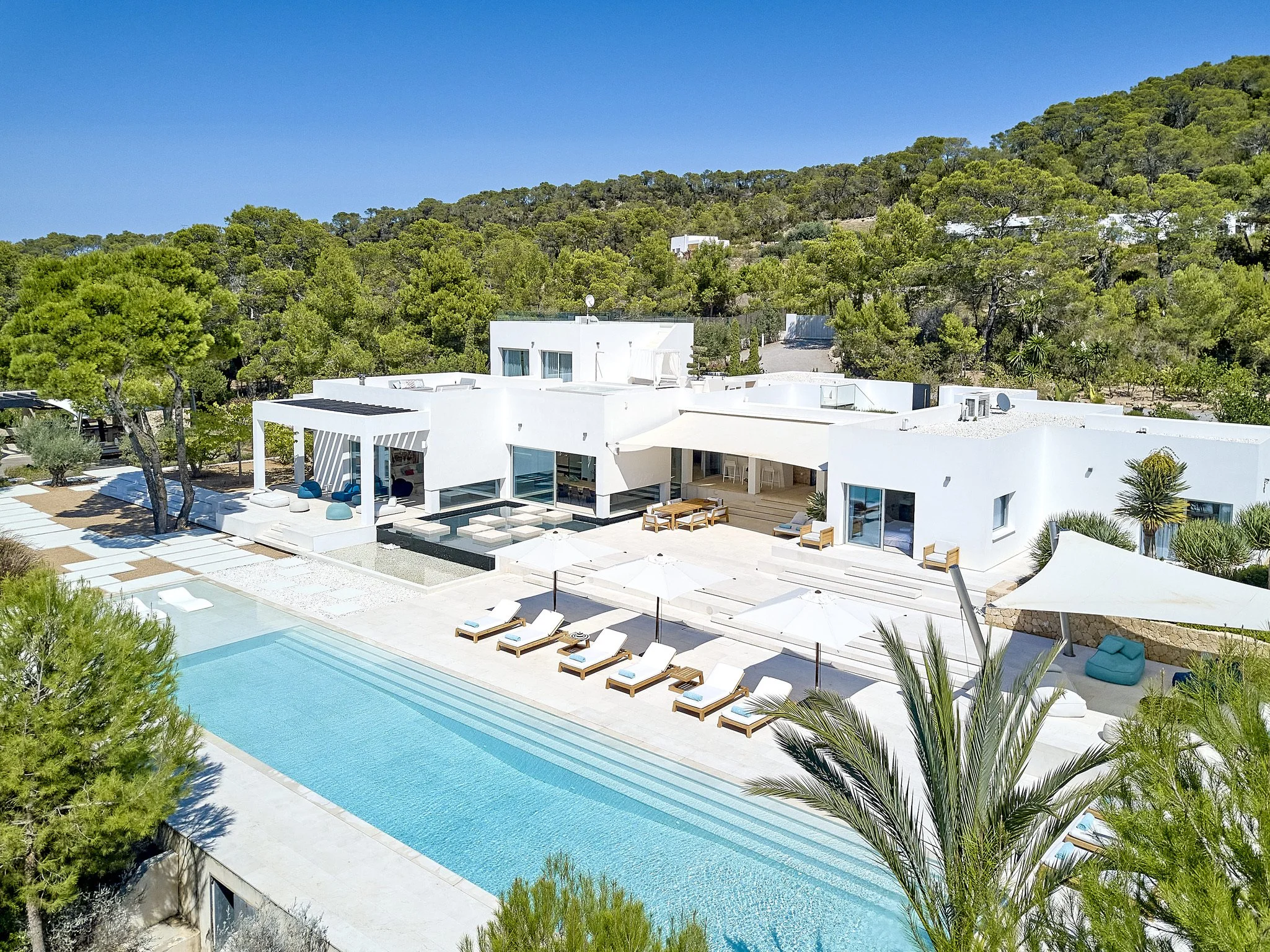 Luxury white villa with a large swimming pool, lounge chairs, outdoor seating, and landscaped grounds surrounded by green hills