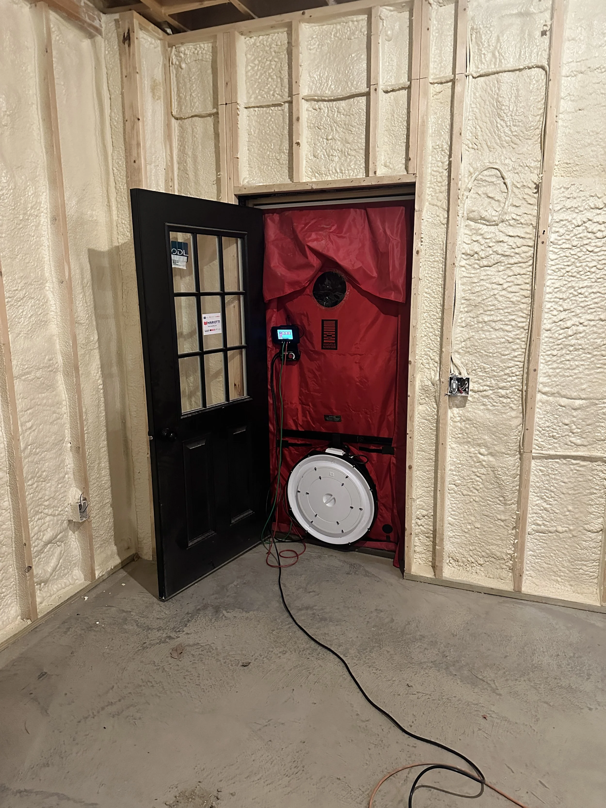 Partially framed basement room with spray-foam insulation, an open black door, and a red temporary dust barrier with an air-testing fan.