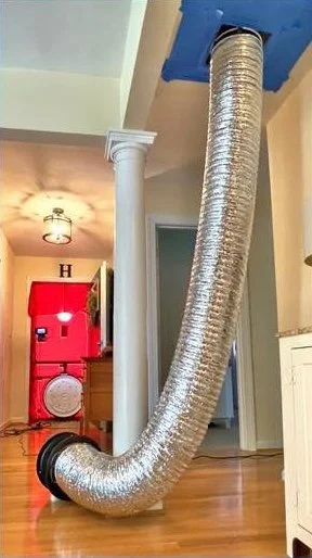 Large flexible silver ventilation duct running from a fan on a hardwood floor to a ceiling opening with a red canvas frame blower door setup in the background.