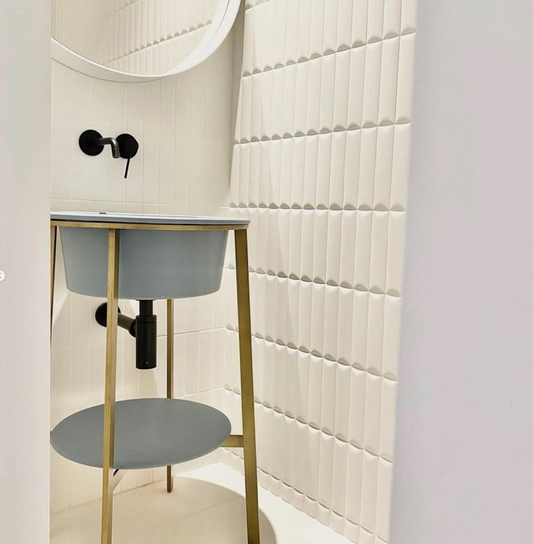 Salle de bains moderne avec lavabo gris sur pieds dorés, robinetterie noire et carrelage blanc texturé.
