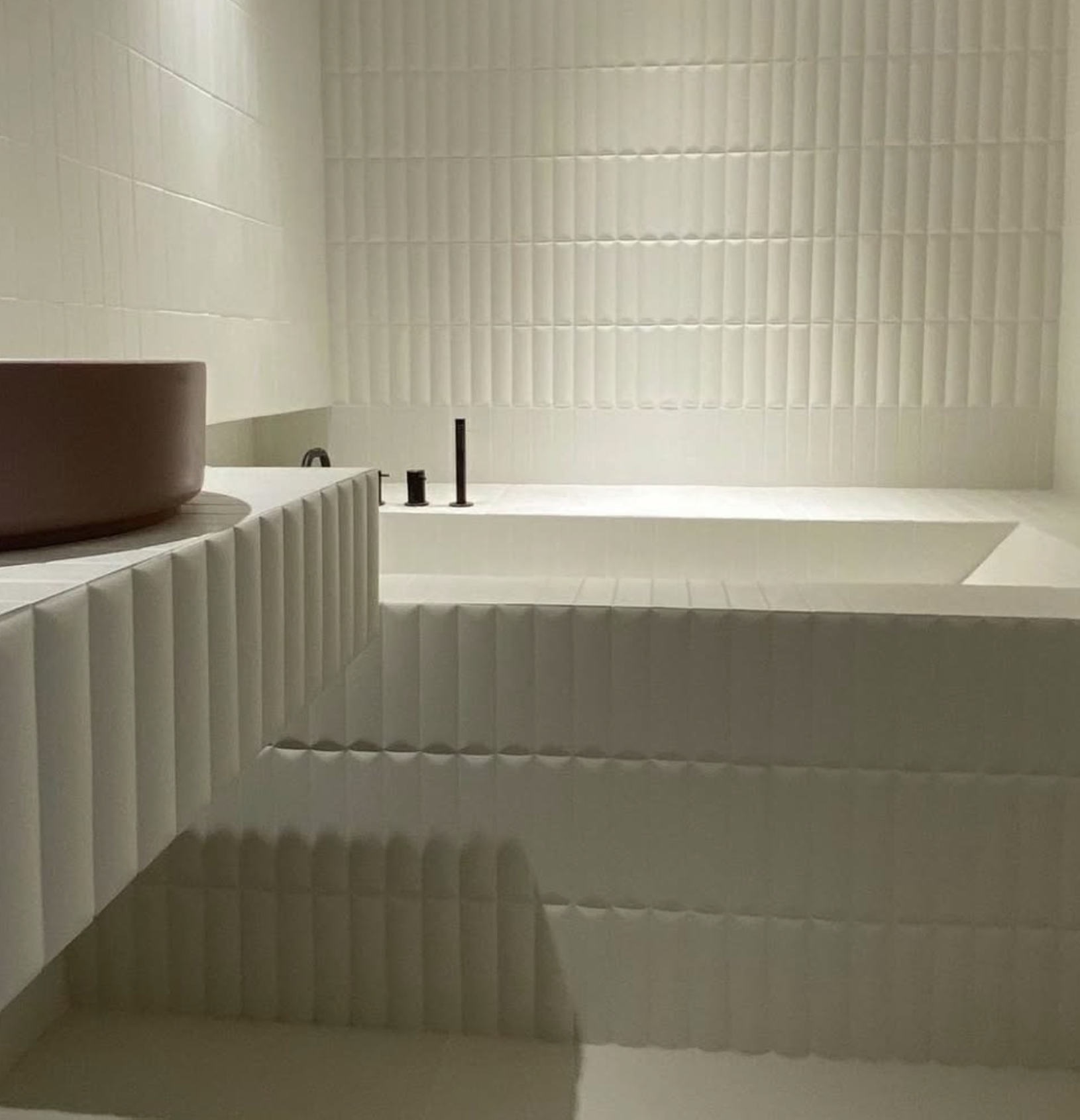 Salle de bains moderne blanche avec baignoire encastrée, lavabo rond et robinetterie noire, recouverte de carrelage texturé.