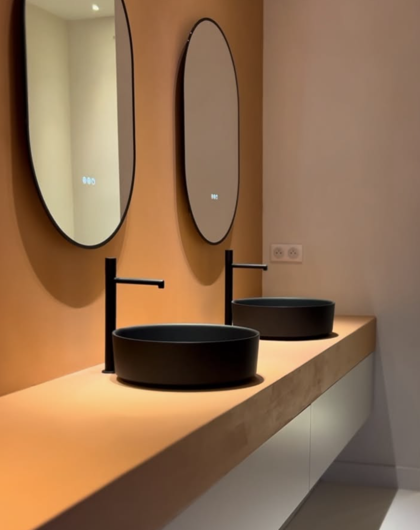 Salle de bains moderne avec deux vasques noires, robinets muraux noirs et miroirs ovales sur un meuble-lavabo en bois.