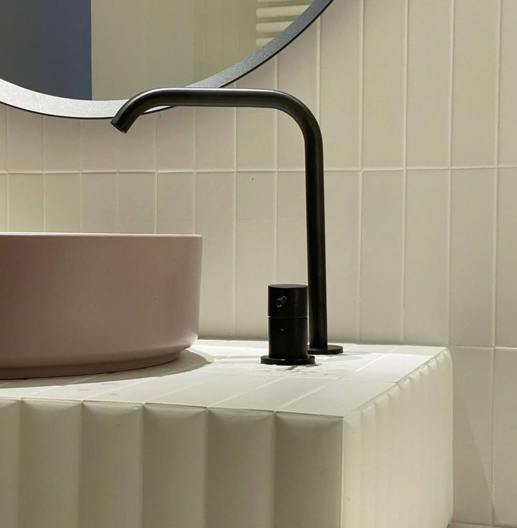 Gros plan sur un robinet noir mat au-dessus d’un lavabo rond beige, dans une salle de bains carrelée de blanc.