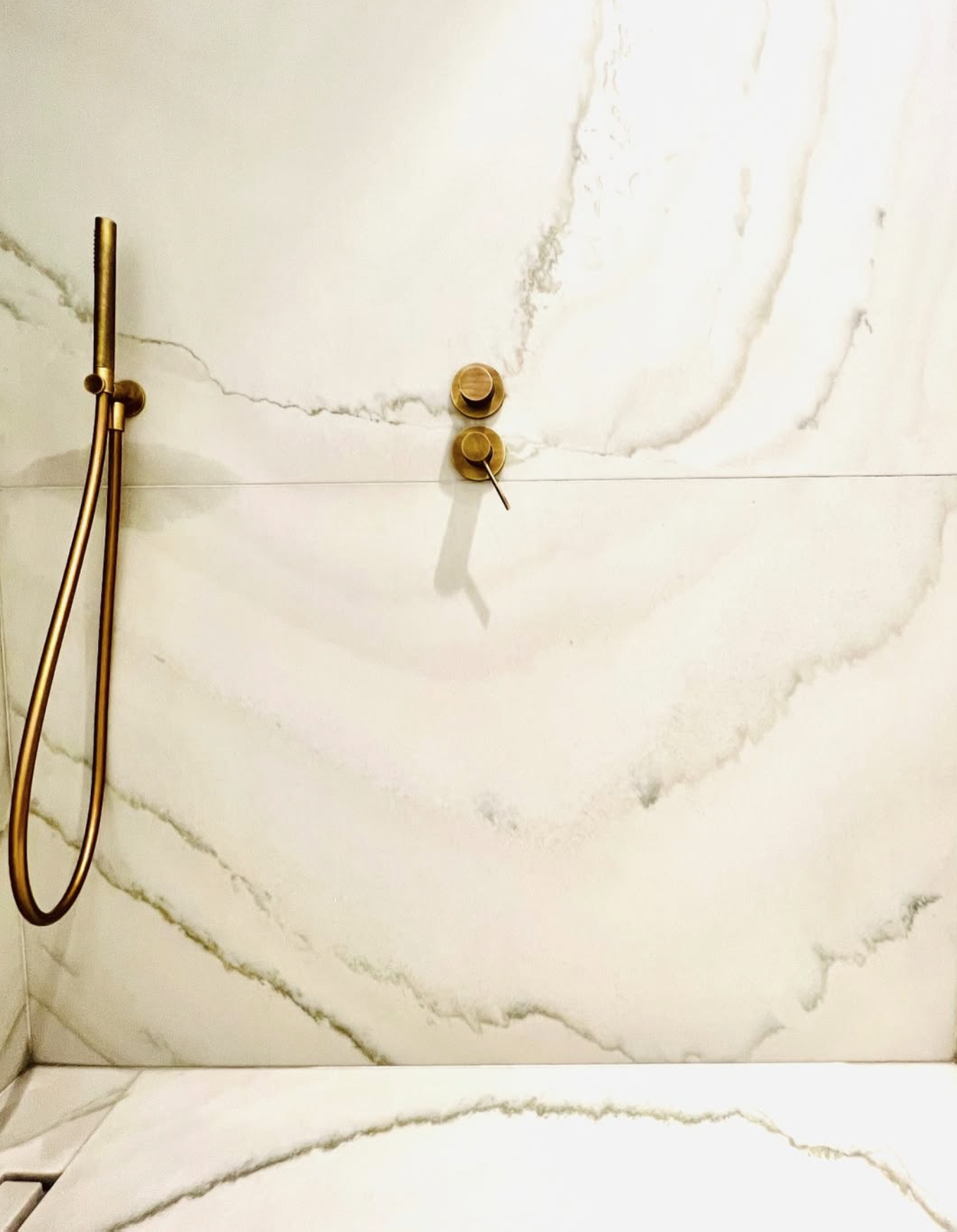 Douche murale en laiton doré fixée sur un carrelage effet marbre blanc, avec commandes rondes et pommeau à main.