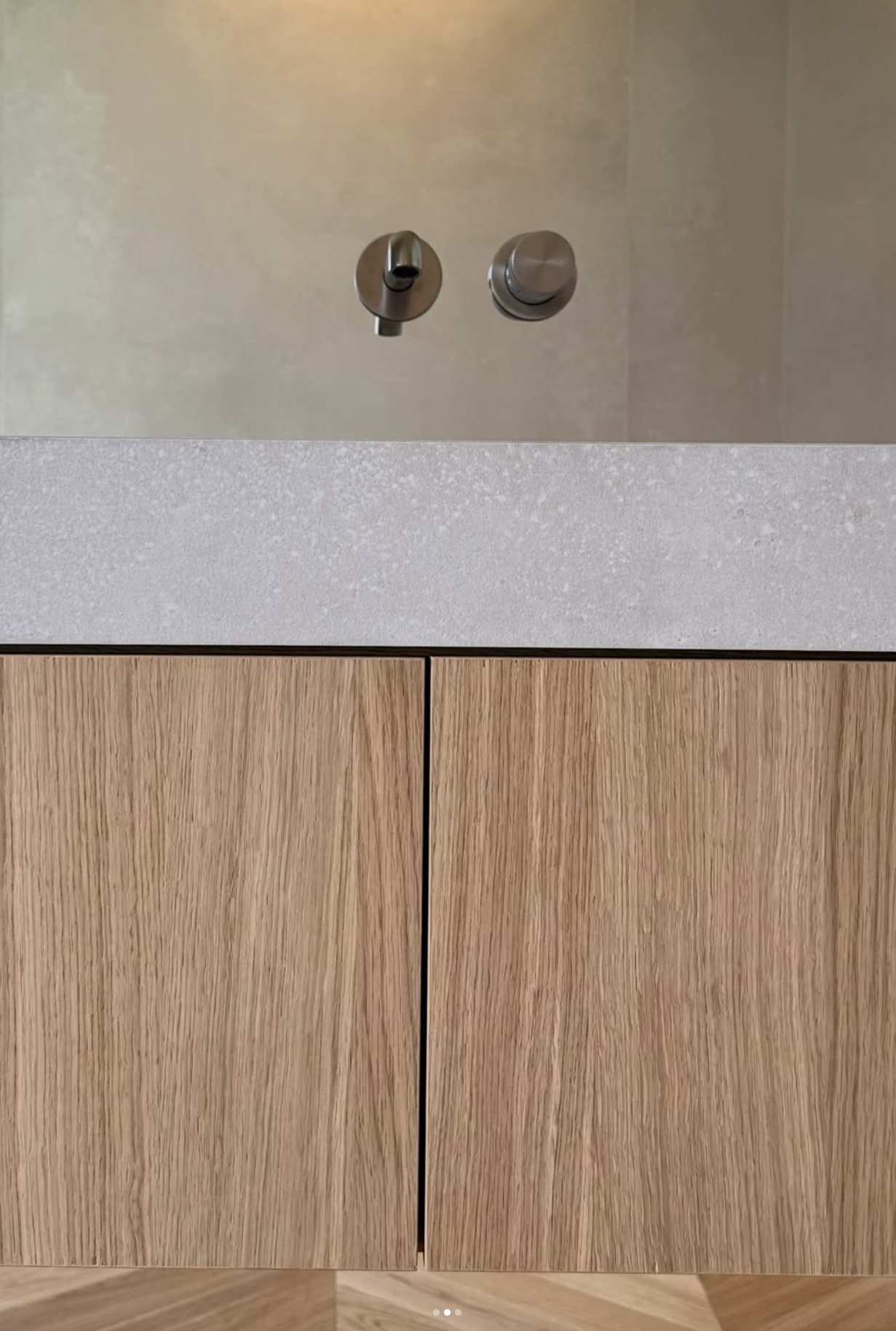 Vue rapprochée d’un meuble-lavabo en bois clair avec deux commandes murales métalliques et un dosseret en pierre grise.