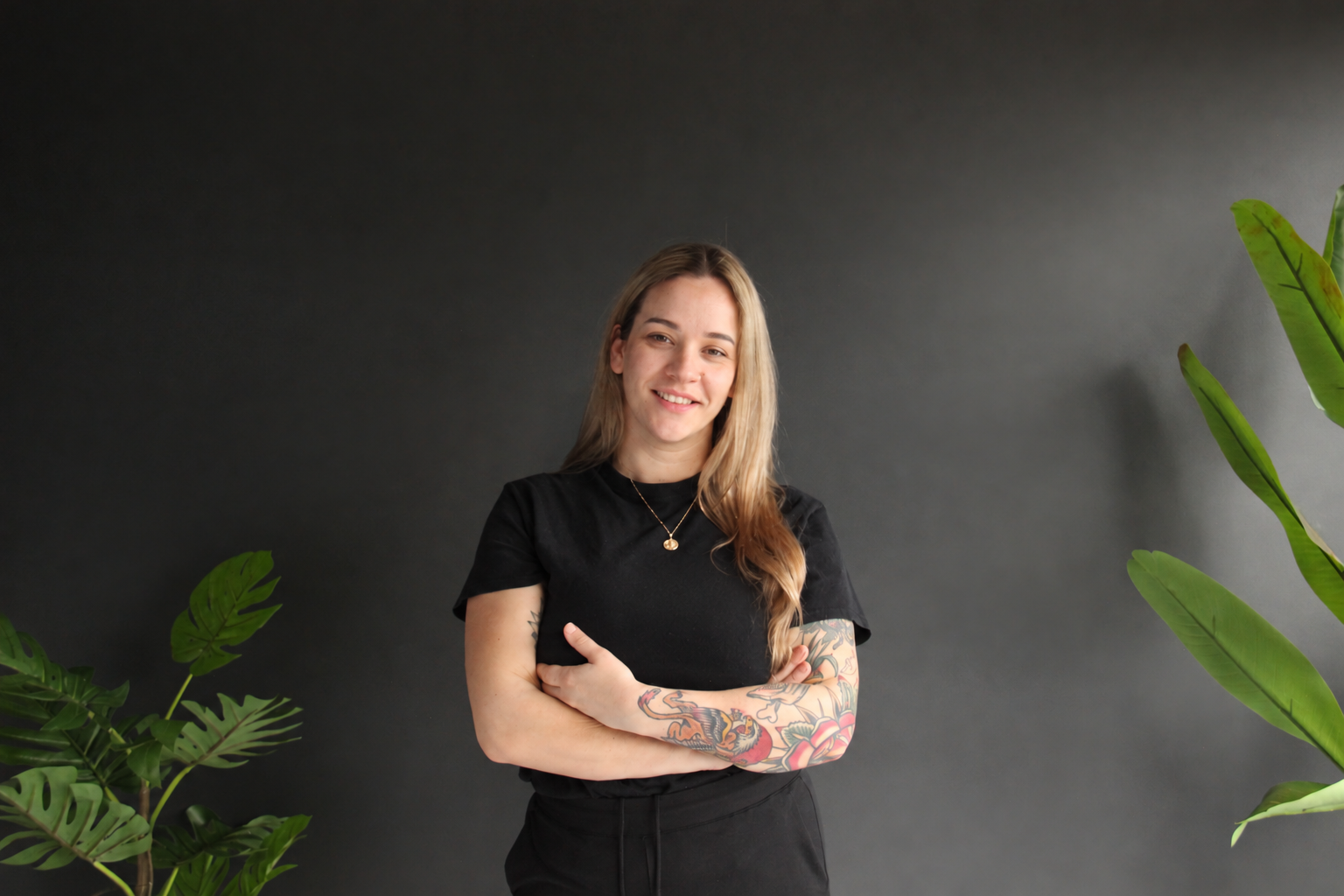 Nicole Wagner, Registered Massage Therapist, smiling with her arms crossed beside green plants.