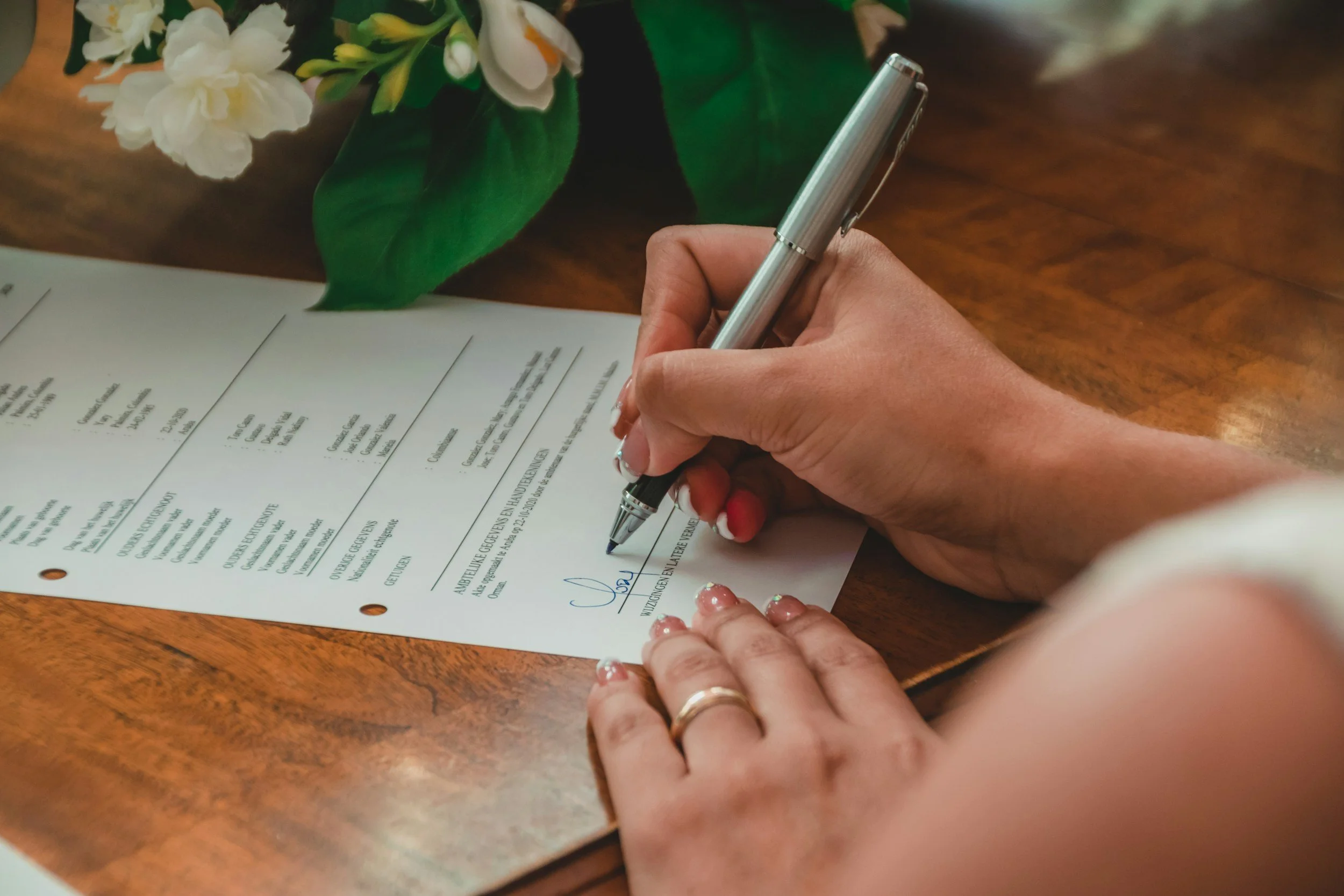 Person signing a printed wedding ceremony document with a silver pen at a wooden table beside white flowers