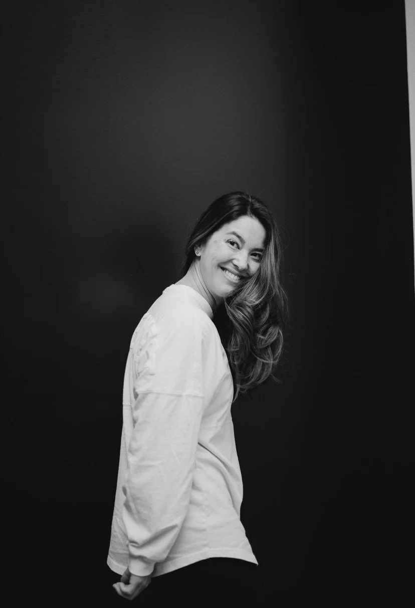 Black-and-white portrait of a smiling woman with long hair wearing a white shirt against a dark background.