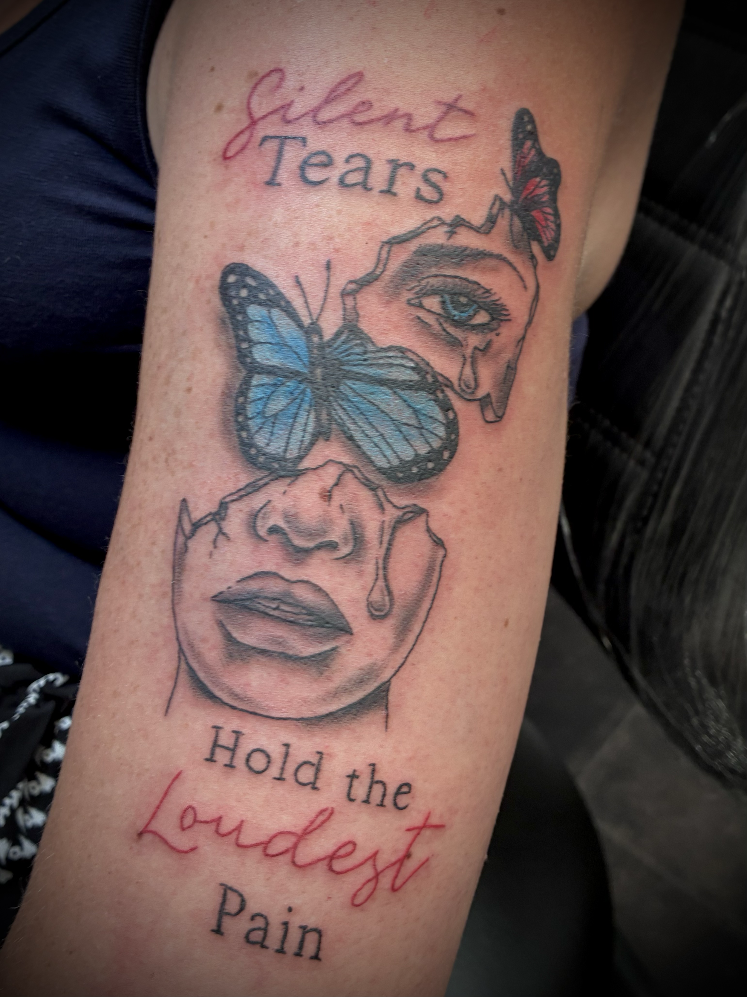 Black and grey tattoo of a woman's face with tears and a blue butterfly, featuring the lettering “Silent Tears, Hold the Loudest Pain”.