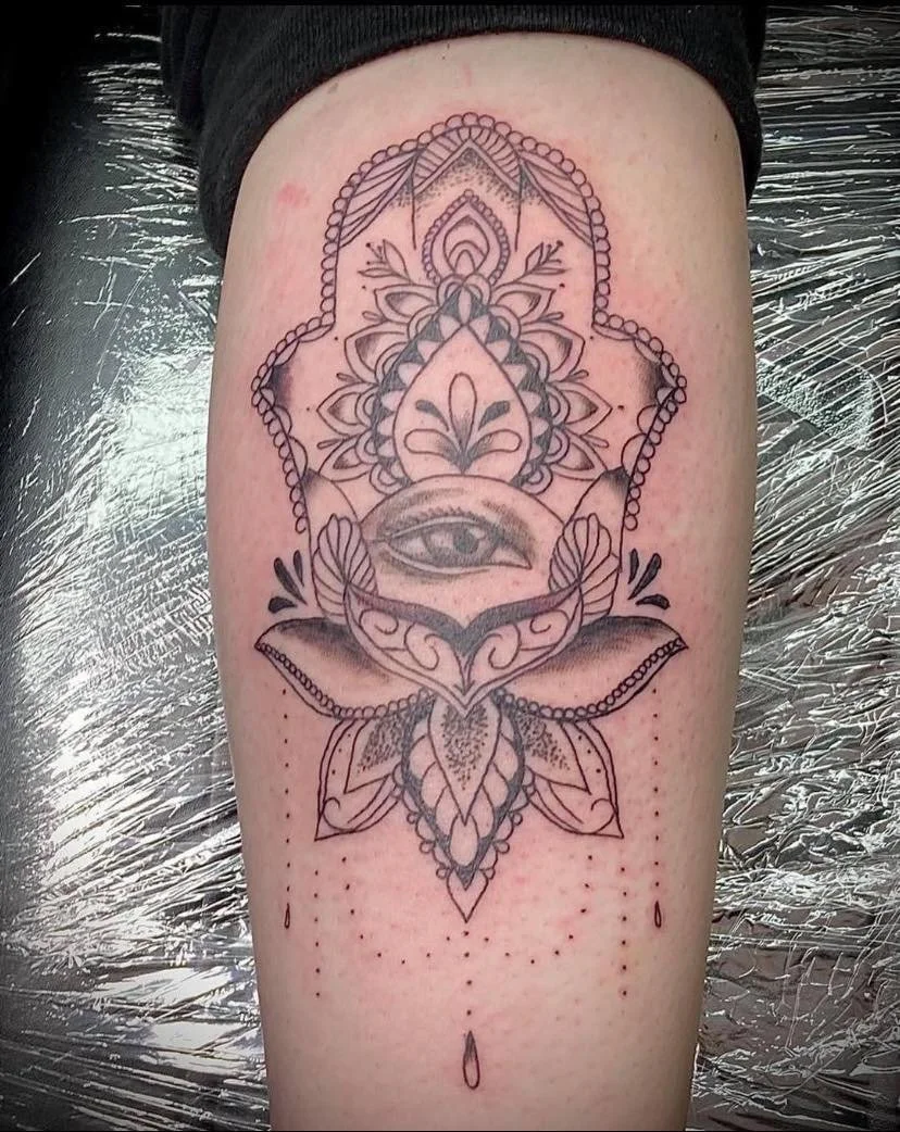 Black and grey ornamental Hamsa hand tattoo featuring a central eye, intricate floral patterns and delicate dotwork shading.