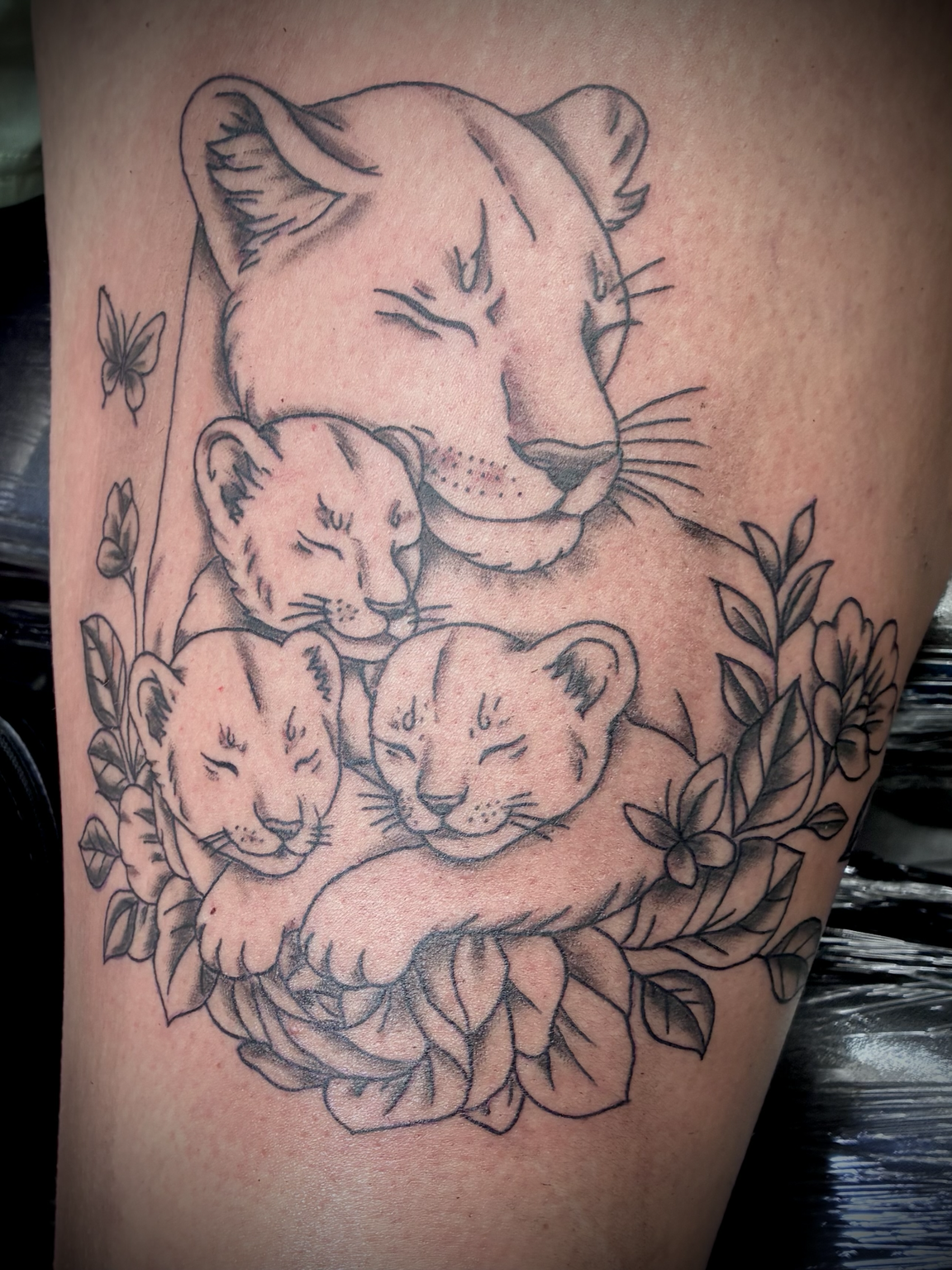 Black and grey fine-line tattoo of a lioness cuddling three lion cubs, surrounded by flowers and leaves.