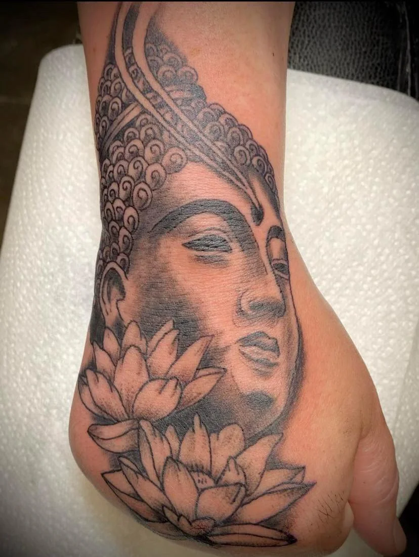 Black and grey Buddha tattoo on the hand and wrist, featuring a peaceful face, intricate ornamental details and lotus flowers.