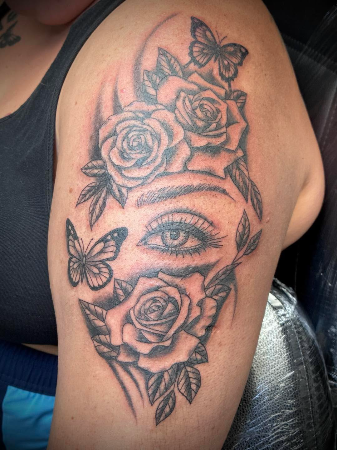Black and grey upper-arm tattoo featuring a realistic eye surrounded by detailed roses, leaves and butterflies.