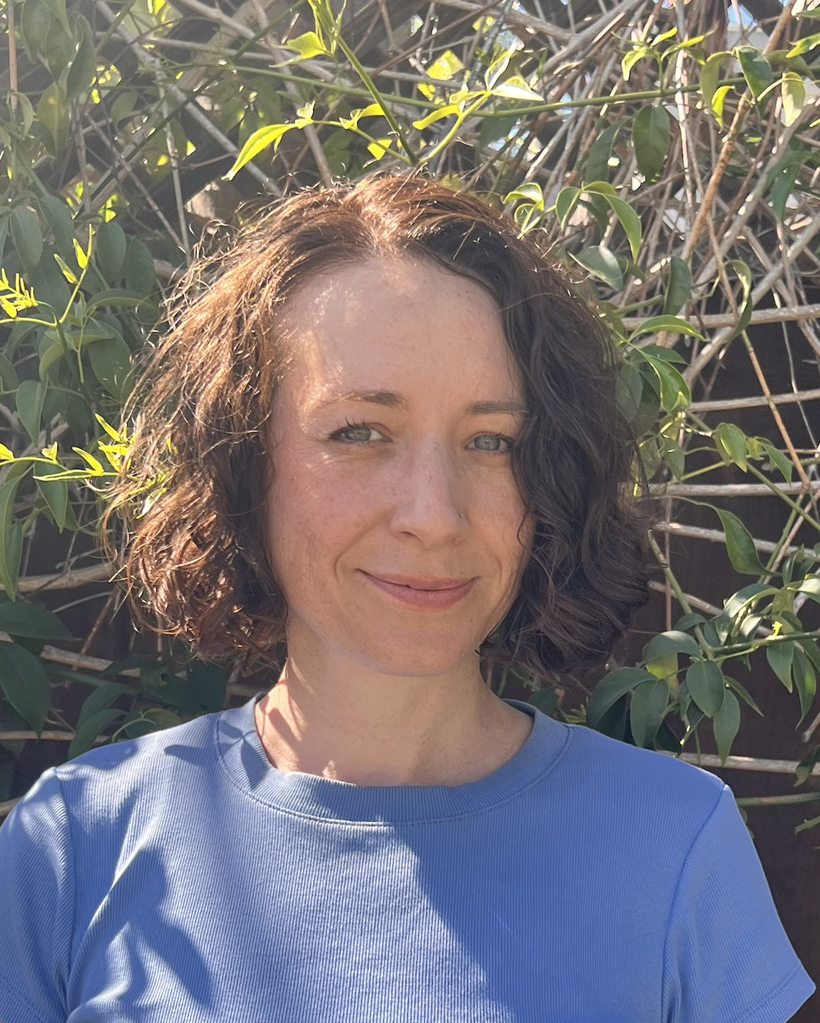 Portrait of a smiling woman with curly brown hair wearing a blue shirt outdoors in front of leafy greenery