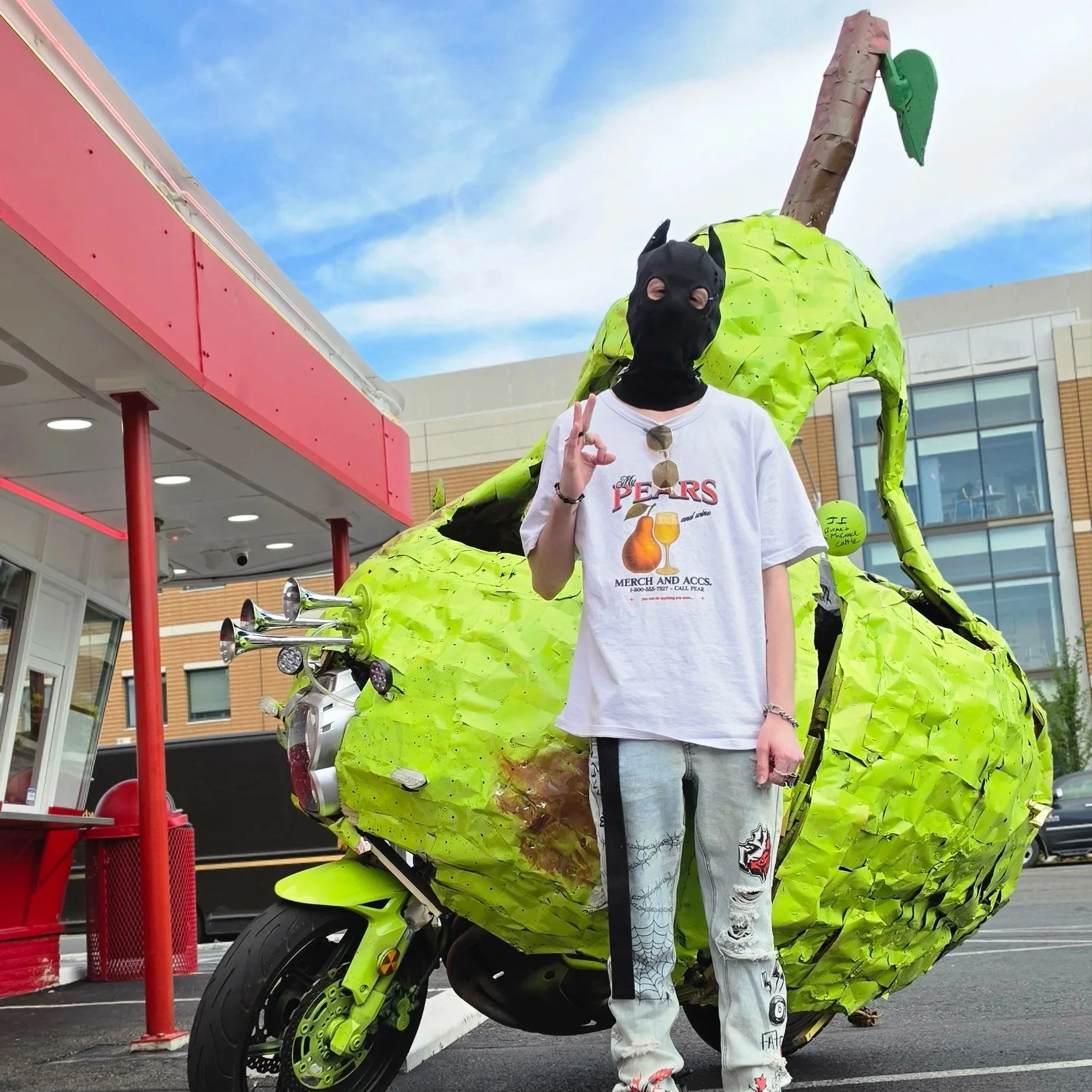 Person wearing a black Batman-style mask and pear-themed T-shirt beside a giant green pear sculpture mounted on a motorcycle outside a building