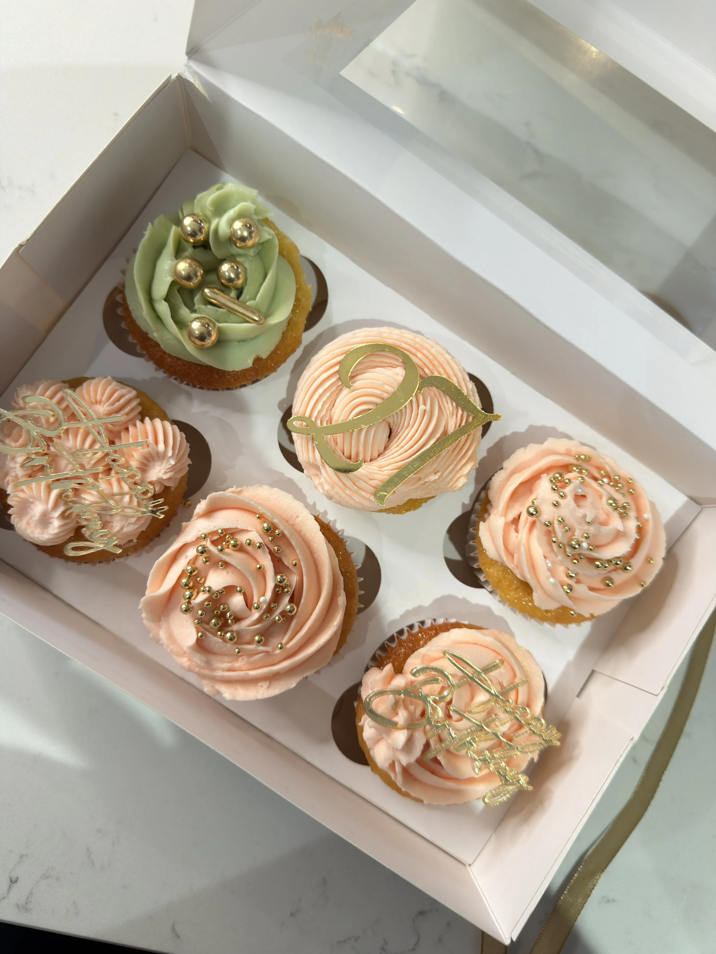 Decorated cupcakes with pastel pink and green frosting, gold sprinkles, and celebratory toppers in a gift box