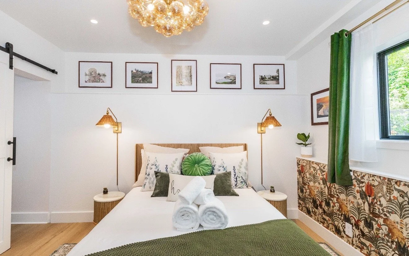 Bright bedroom with a neatly made bed, rolled towels, green accents, framed artwork, wall sconces, and a floral upholstered bench by the window