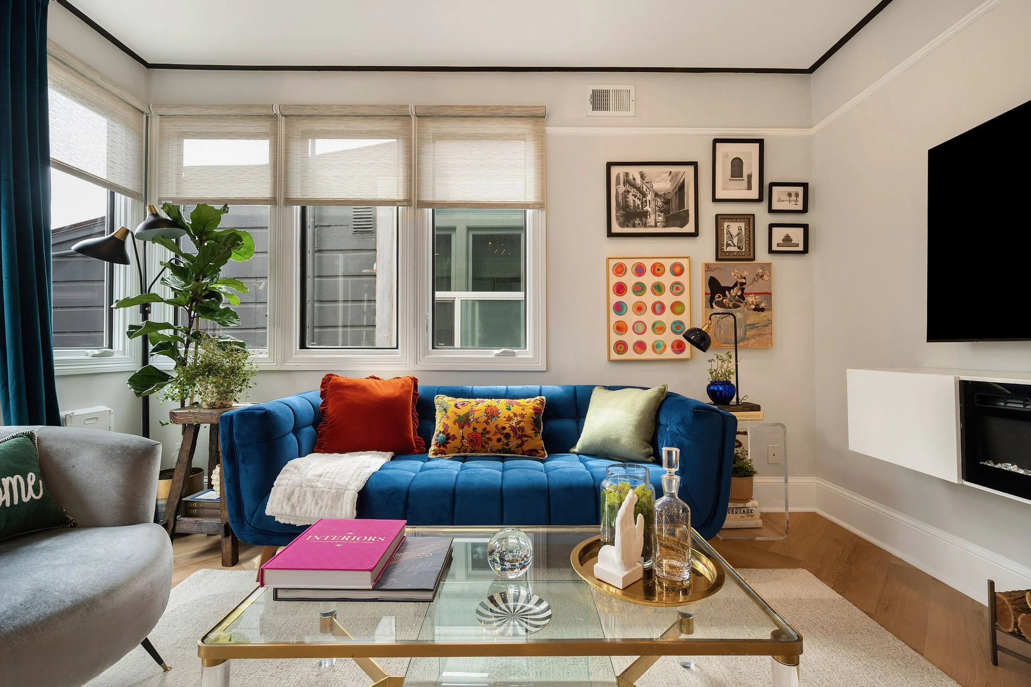 Bright living room with a blue velvet sofa, colorful pillows, indoor plants, framed artwork, large windows, glass coffee table, and wall-mounted TV