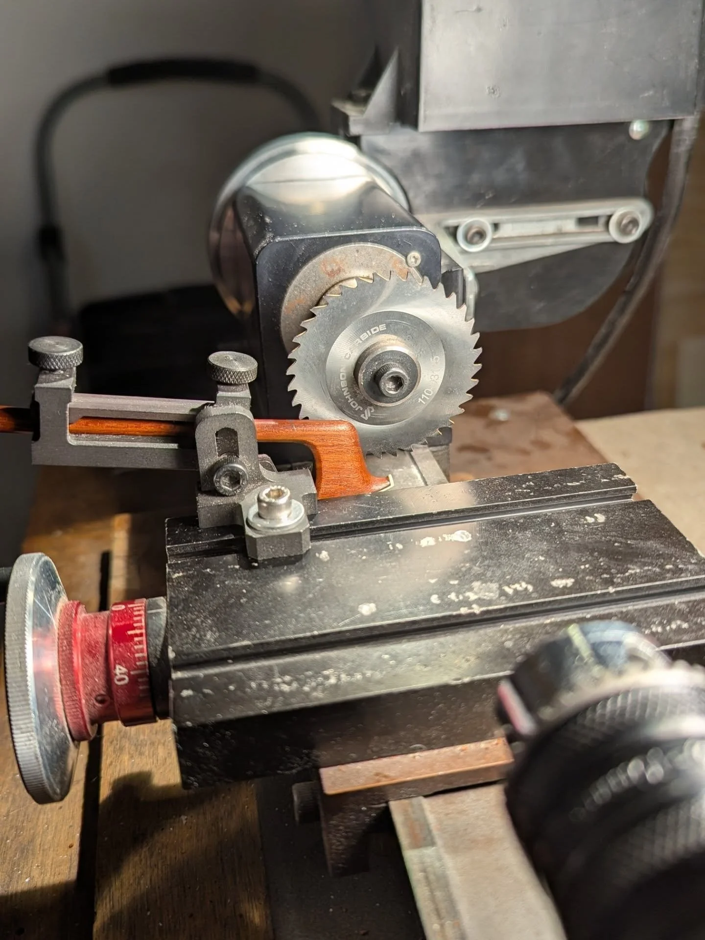 Close-up of a benchtop woodworking machine with a circular saw blade, violin bow, adjustable guides, and a black worktable.