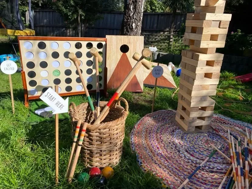 Outdoor carnival game display with Connect Four, giant wooden Jenga tower, croquet mallets, colorful balls, and signs on a grassy lawn