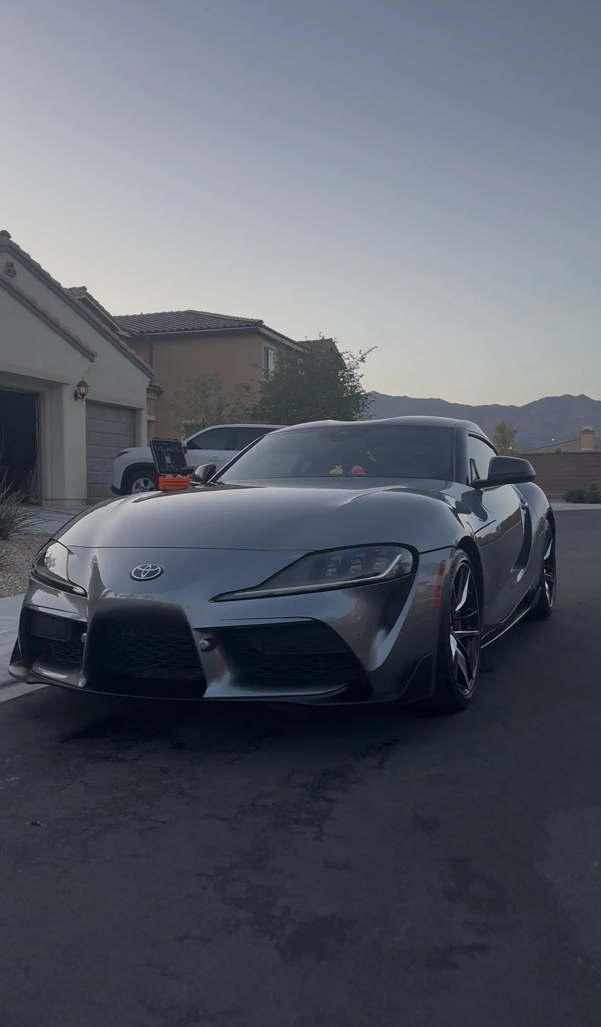 Gray Toyota Supra sports car parked in a suburban driveway at dusk with mountains in the background
