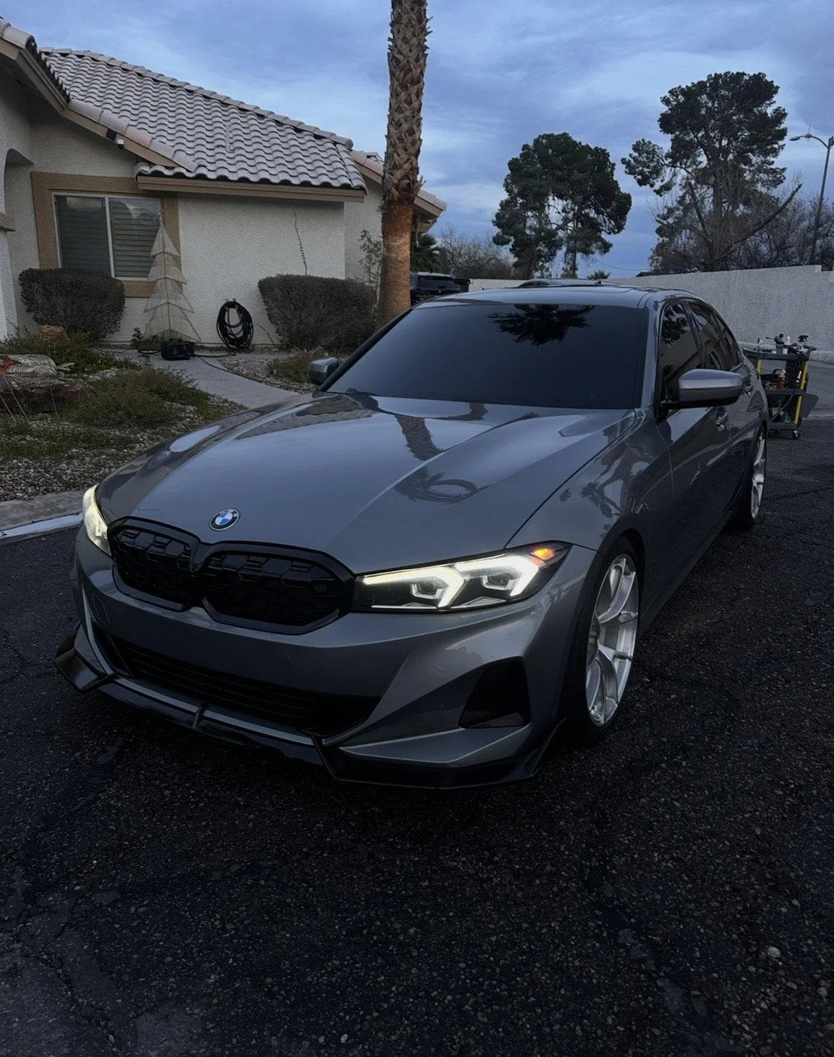 Gray BMW sedan with tinted windows and aftermarket wheels parked in a residential driveway at dusk