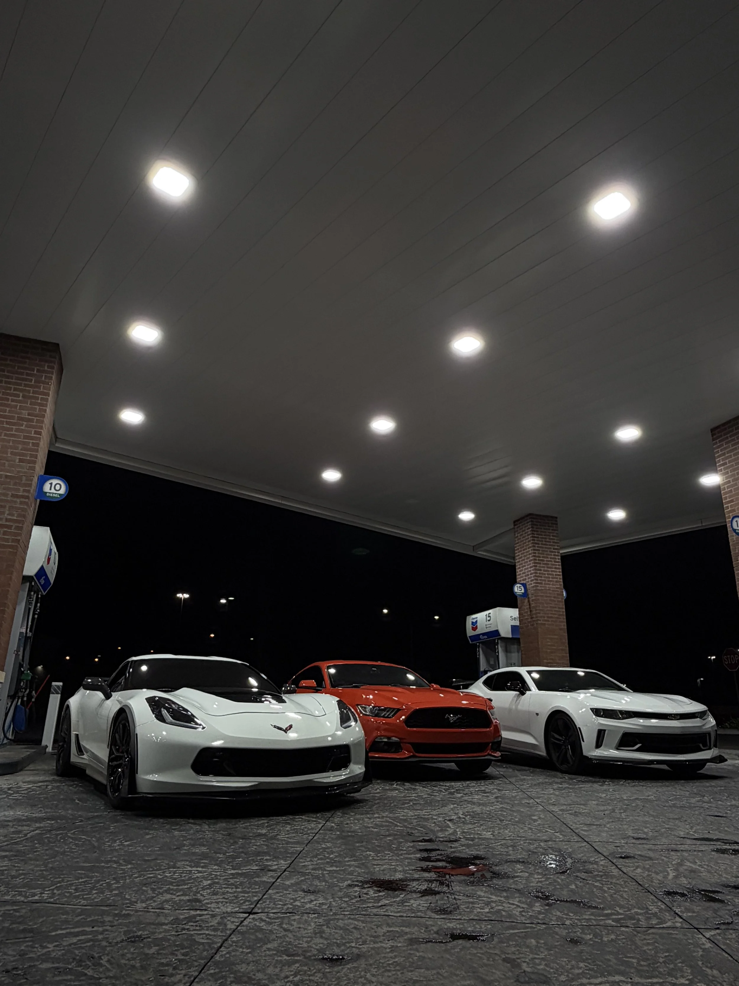 Three sports cars—a white Chevrolet Corvette, red Ford Mustang, and white Chevrolet Camaro—parked at a brightly lit gas station at night.