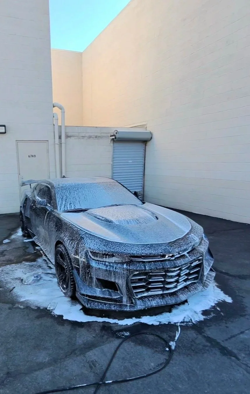 Black Chevrolet Camaro covered in soap suds at a car wash outside a commercial building