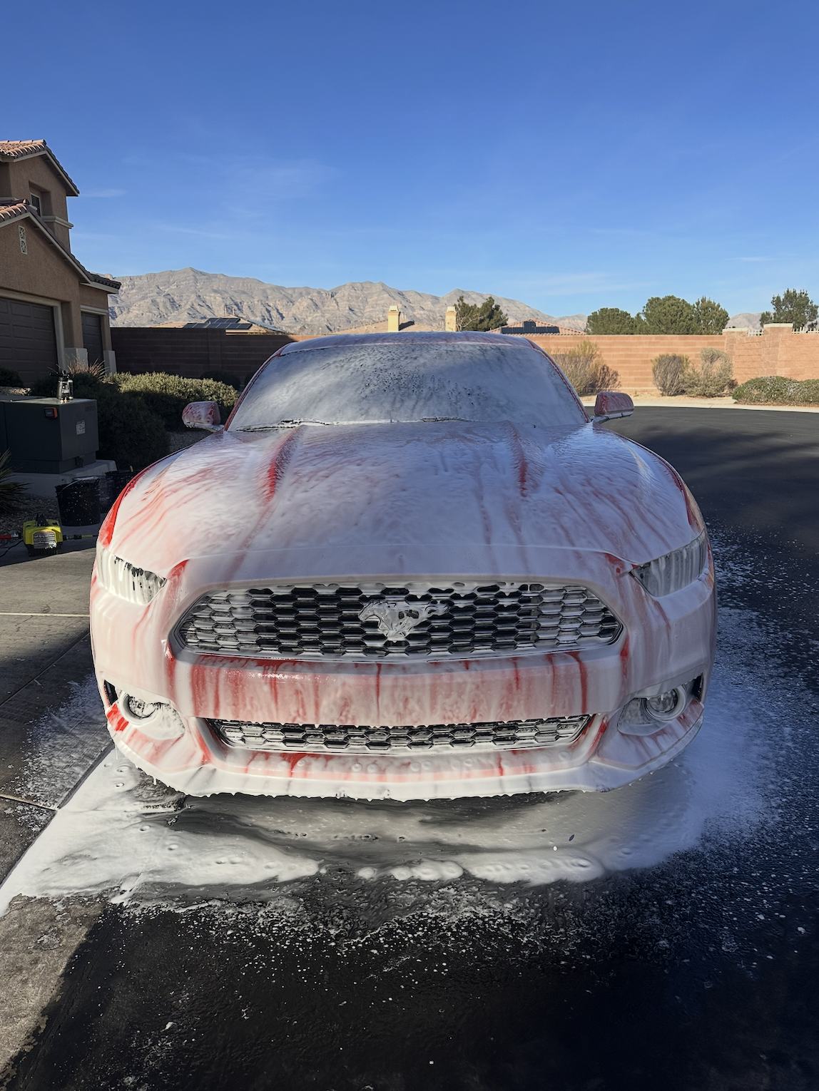 Front view of a Ford Mustang covered in white and red car wash foam in a driveway, with mountains in the background