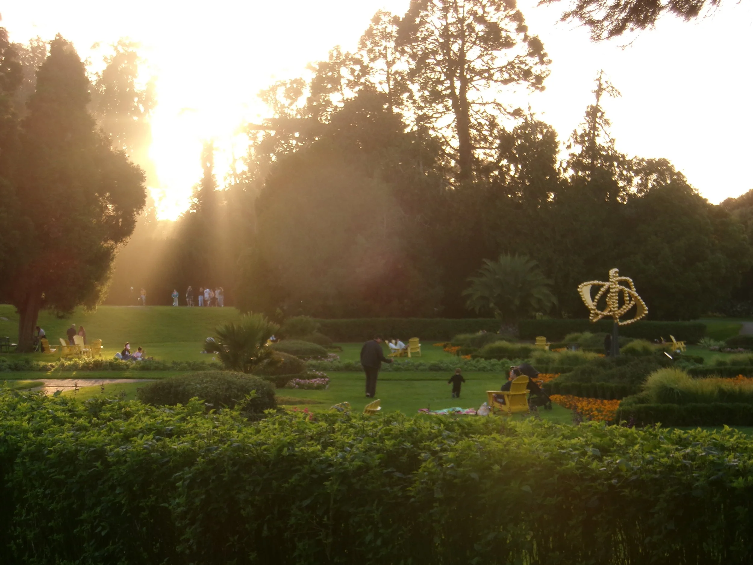 People relaxing in a landscaped garden at sunset, surrounded by flower beds, trees, and a crown-shaped illuminated sculpture