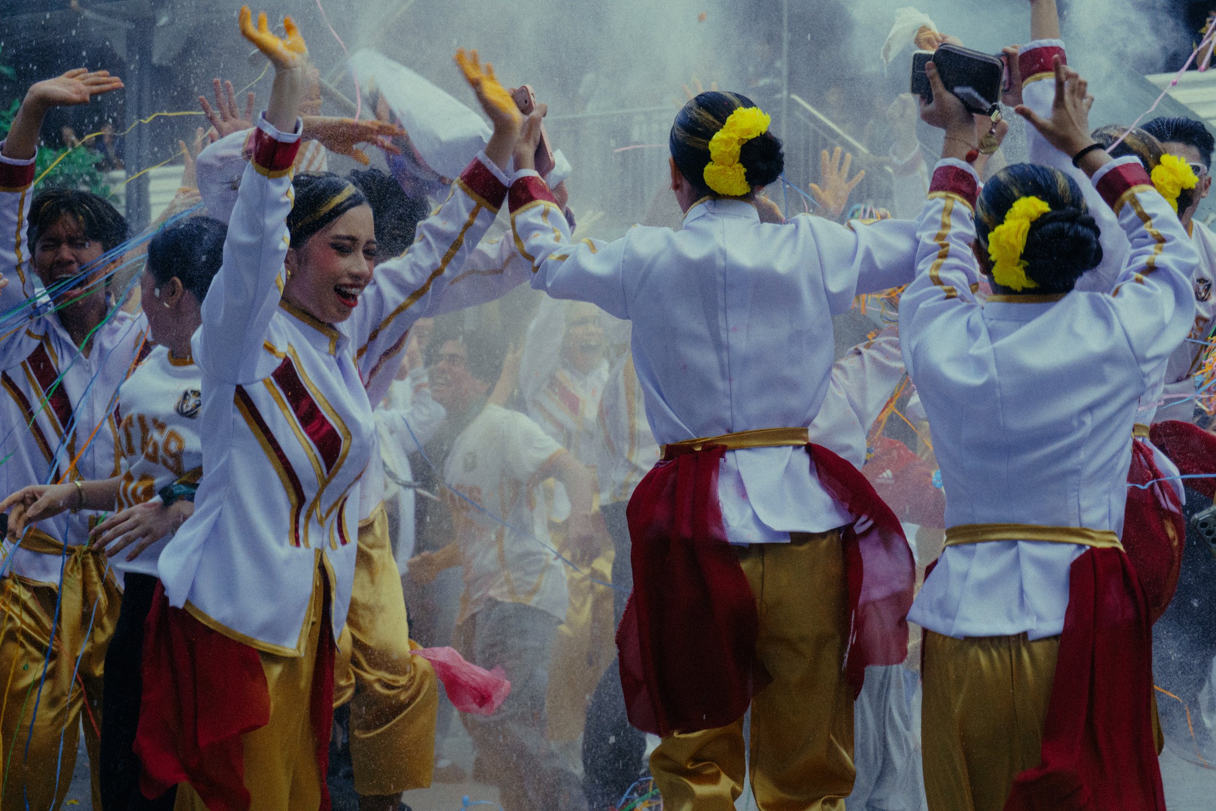Crowd in colorful white, red, and gold costumes dancing amid confetti, with yellow flowers in their hair and people recording on smartphones.
