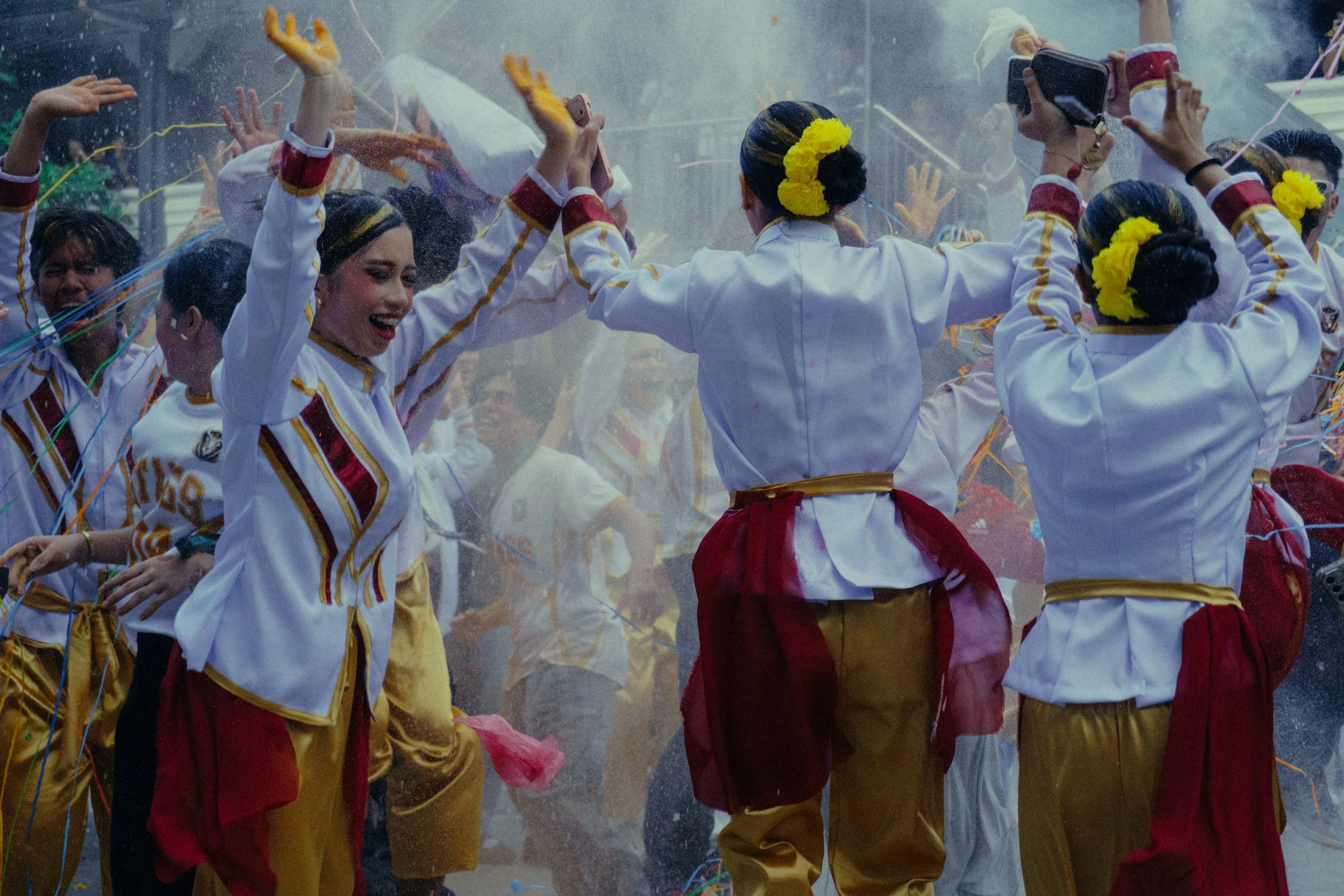 Crowd in colorful white, red, and gold costumes dancing amid confetti, with yellow flowers in their hair.
